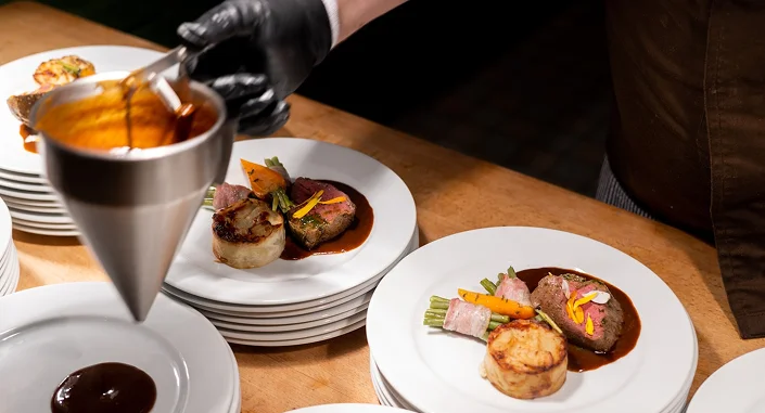Chef-kok die zorgvuldig de opgemaakte hoofdgerechten afwerkt met vlees, groenten en rijke saus in een professionele restaurantkeuken.